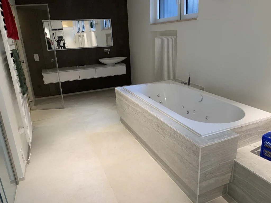 Badezimmer mit Whirlpool-Wanne, Wannenverkleidung aus hell gestreiftem Naturstein und großformatigen hellen Bodenfliesen