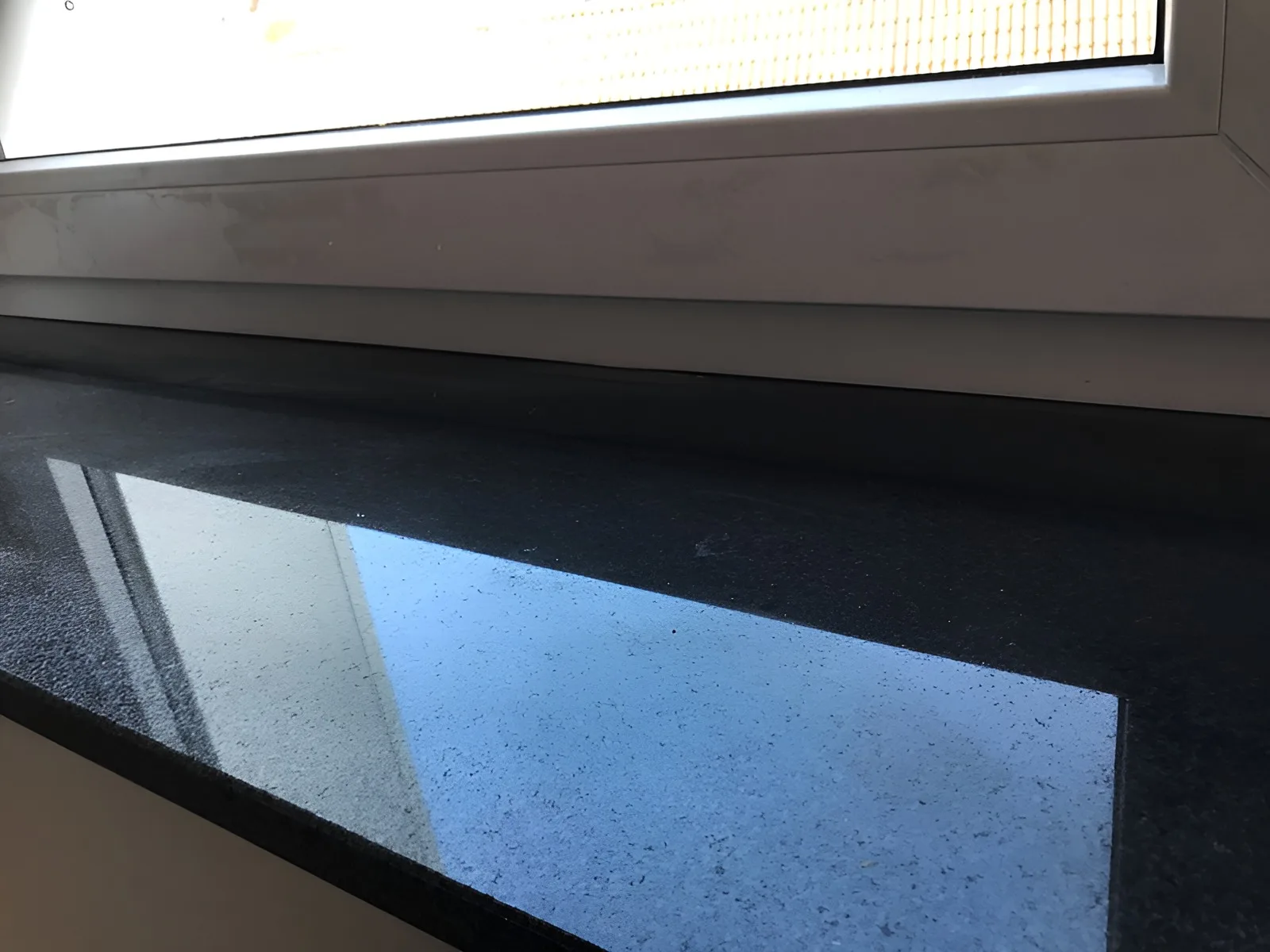 Polierte Fensterbank aus schwarzem Granit unter einem weißen Kunststofffenster, das Fenster spiegelt sich in der Oberfläche