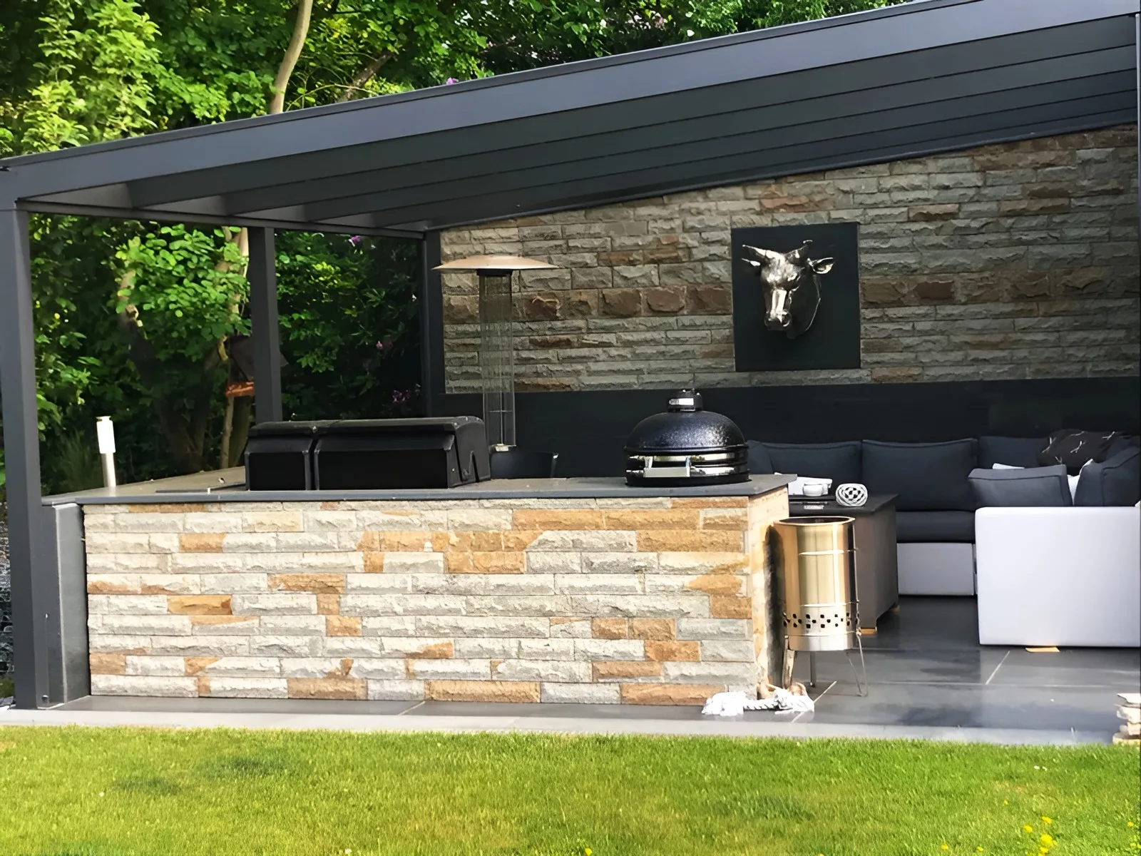 Überdachte Außenküche mit Grill und Lounge-Ecke, Theke und Rückwand mit hellen Natursteinriemchen verblendet, Boden aus dunklen Platten